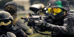 En medio de una intensa partida de Mallorca Paintball, un jugador equipado de forma completa apunta su arma con precisión, mientras sus compañeros elaboran estrategias con el telón de fondo de los 5 Campos Tematizados. - ForTwo - Planes para parejas