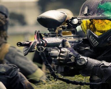 En medio de una intensa partida de Mallorca Paintball, un jugador equipado de forma completa apunta su arma con precisión, mientras sus compañeros elaboran estrategias con el telón de fondo de los 5 Campos Tematizados. - ForTwo - Planes para parejas