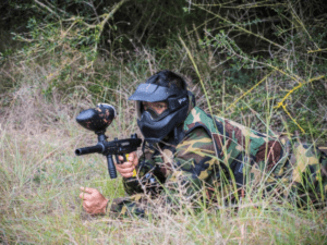 Vestido de camuflaje y con una máscara, un jugador se mueve hábilmente por el terreno en Mallorca Paintball: 5 Campos Tematizados, tumbado en el césped mientras apunta su pistola de paintball para vivir una emocionante aventura. - ForTwo - Planes para parejas
