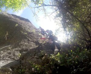 Utilizando cuerdas, los aventureros escalan un acantilado rocoso y escarpado rodeado de exuberante vegetación y un cielo azul claro en la experiencia Multiaventura en el Barranco de la Luna. - ForTwo - Planes para parejas