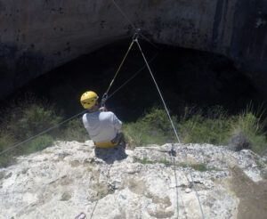 Con un casco y asegurado con cuerdas, alguien se sienta en el borde del acantilado con vistas a la entrada de la cueva del Barranco de la Luna, preparado para vivir una experiencia emocionante con Multiaventura en el Barranco de la Luna. - ForTwo - Planes para parejas