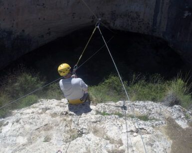 Con un casco y asegurado con cuerdas, alguien se sienta en el borde del acantilado con vistas a la entrada de la cueva del Barranco de la Luna, preparado para vivir una experiencia emocionante con Multiaventura en el Barranco de la Luna. - ForTwo - Planes para parejas