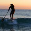Una persona disfruta de "Padel surf: Remo al Atardecer en Fuengirola" al atardecer, deslizándose sobre una pequeña ola mientras el océano sereno refleja el cielo naranja vibrante. - ForTwo - Planes para parejas