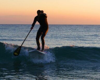 Una persona disfruta de "Padel surf: Remo al Atardecer en Fuengirola" al atardecer, deslizándose sobre una pequeña ola mientras el océano sereno refleja el cielo naranja vibrante. - ForTwo - Planes para parejas