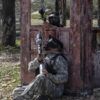 Dos personas con equipo de camuflaje y máscaras disfrutan de un emocionante juego de paintball detrás de una estructura de madera desgastada, similar a "Paintball a 10 minutos de Mallorca: Aventura y Diversión. - ForTwo - Planes para parejas