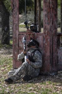 Dos personas con equipo de camuflaje y máscaras disfrutan de un emocionante juego de paintball detrás de una estructura de madera desgastada, similar a "Paintball a 10 minutos de Mallorca: Aventura y Diversión. - ForTwo - Planes para parejas
