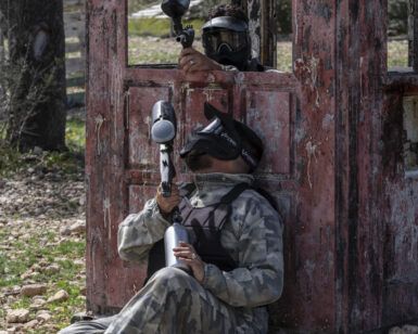 Dos personas con equipo de camuflaje y máscaras disfrutan de un emocionante juego de paintball detrás de una estructura de madera desgastada, similar a "Paintball a 10 minutos de Mallorca: Aventura y Diversión. - ForTwo - Planes para parejas