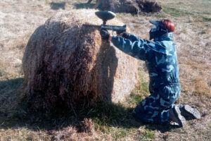 En Jaca, un jugador con traje de camuflaje se arrodilla junto a un fardo de heno y apunta con precisión su pistola de paintball. "Paintball en Jaca: Diversión y Estrategia en Escenarios de Alto Nivel" transforma el campo en un emocionante campo de batalla. - ForTwo - Planes para parejas