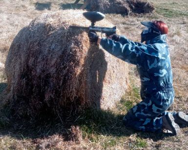 En Jaca, un jugador con traje de camuflaje se arrodilla junto a un fardo de heno y apunta con precisión su pistola de paintball. "Paintball en Jaca: Diversión y Estrategia en Escenarios de Alto Nivel" transforma el campo en un emocionante campo de batalla. - ForTwo - Planes para parejas