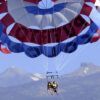 Dos amantes de las emociones disfrutan de "Parascending en Costa Adeje: Vuela y Disfruta de las Vistas del Sur de Tenerife", con un paracaídas rojo, blanco y azul frente al impresionante telón de fondo montañoso de Costa Adeje en Tenerife. - ForTwo - Planes para parejas