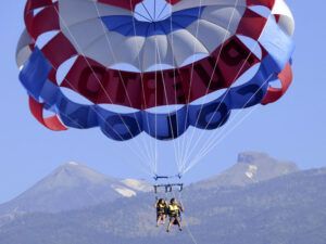 Dos amantes de las emociones disfrutan de "Parascending en Costa Adeje: Vuela y Disfruta de las Vistas del Sur de Tenerife", con un paracaídas rojo, blanco y azul frente al impresionante telón de fondo montañoso de Costa Adeje en Tenerife. - ForTwo - Planes para parejas