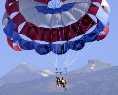 Dos amantes de las emociones disfrutan de "Parascending en Costa Adeje: Vuela y Disfruta de las Vistas del Sur de Tenerife", con un paracaídas rojo, blanco y azul frente al impresionante telón de fondo montañoso de Costa Adeje en Tenerife. - ForTwo - Planes para parejas