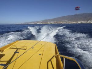 En la experiencia Parascending en Costa Adeje, disfrute de las vistas desde un barco amarillo deslizándose por el océano cerca de Costa Adeje en Tenerife, con un vibrante parasail en el cielo y majestuosas montañas en el horizonte. - ForTwo - Planes para parejas
