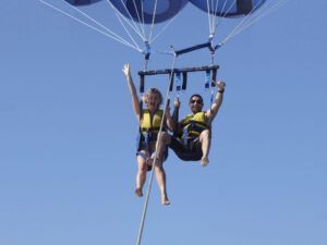 Dos personas disfrutan de un descenso en paracaídas sobre el claro cielo azul de Costa Adeje, usando chalecos salvavidas y sonriendo mientras levantan los brazos con alegría. Están viviendo la emoción del "Parascending en Costa Adeje: Vuela y Disfruta de las Vistas del Sur de Tenerife. - ForTwo - Planes para parejas
