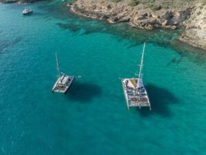 Un "Paseo en Barco al Atardecer en Mallorca: Sunset Tour" presenta dos veleros deslizándose por las cristalinas aguas turquesas de Mallorca y un pequeño barco a la deriva en la distancia cerca de la costa rocosa. - ForTwo - Planes para parejas