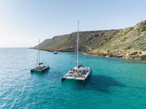 Dos catamaranes navegan en aguas cristalinas color turquesa cerca de una costa rocosa bajo un cielo azul vibrante, capturando la esencia de Paseo en Barco al Atardecer en Mallorca: Tour al Atardecer. Experimente la serena belleza mientras el atardecer proyecta su resplandor dorado sobre el pintoresco paisaje marino. - ForTwo - Planes para parejas