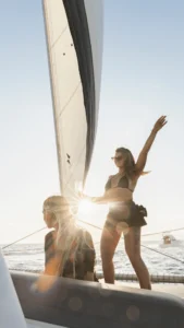 Dos mujeres disfrutan de un "Paseo en Barco al Atardecer en Mallorca: Sunset Tour"; una permanece de pie con los brazos en alto en señal de júbilo mientras la otra se sienta serenamente en el velero bajo el sol dorado, con un océano infinito como telón de fondo. - ForTwo - Planes para parejas