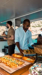 Un hombre con gafas de sol sirve comida tipo buffet en un barco, mientras la gente disfruta de la vista durante el Paseo en Barco al Atardecer en Mallorca: Sunset Tour, añadiendo magia a las brillantes aguas nocturnas de Mallorca. - ForTwo - Planes para parejas