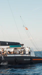 Un catamarán surca el océano bajo un cielo despejado frente a la costa de Mallorca, con una colorida bandera ondeando en el mástil. Los pasajeros disfrutan del Paseo en Barco al Atardecer en Mallorca: Tour al Atardecer, deleitándose con la belleza del atardecer. - ForTwo - Planes para parejas