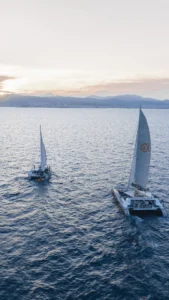 Durante el Paseo en Barco al Atardecer en Mallorca: Sunset Tour, dos veleros se deslizan graciosamente sobre aguas tranquilas con un telón de fondo de montañas lejanas y un cielo pálido. - ForTwo - Planes para parejas