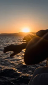 Silueta de manos y brazos en un barco durante el Paseo en Barco al Atardecer en Mallorca: Sunset Tour, con el sol poniéndose sobre montañas lejanas y mar en calma. - ForTwo - Planes para parejas