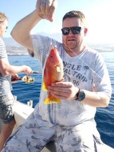 Un hombre en un barco frente a la costa de Tenerife sostiene con orgullo un pez vibrante capturado durante su aventura de Pesca Deportiva en Tenerife con SEA PASSION, mientras otro disfruta del deporte en el fondo. - ForTwo - Planes para parejas
