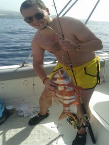 Un hombre con pantalones cortos amarillos, mostrando su amor por la pesca, sostiene con orgullo una caña y un pez grande mientras está a bordo de su barco frente a la costa de Tenerife durante Pesca Deportiva en Tenerife con SEA PASSION. - ForTwo - Planes para parejas