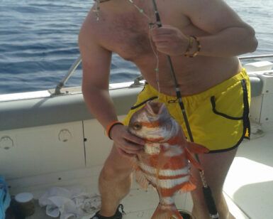 Un hombre con pantalones cortos amarillos, mostrando su amor por la pesca, sostiene con orgullo una caña y un pez grande mientras está a bordo de su barco frente a la costa de Tenerife durante Pesca Deportiva en Tenerife con SEA PASSION. - ForTwo - Planes para parejas