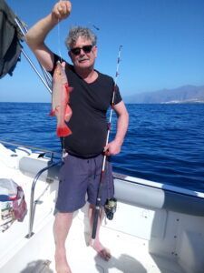 En un barco en Tenerife, un hombre exhibe con orgullo su captura, un pez rojo, con su caña de pescar en alto. Esta escena encarna la esencia de la Pesca Deportiva en Tenerife con PASIÓN MARINA, con el mar azul de fondo. - ForTwo - Planes para parejas