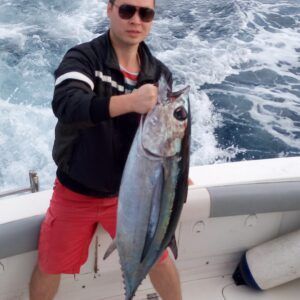 Una persona con gafas de sol sostiene con orgullo un gran pez en un barco, encarnando la "Pesca Deportiva en Tenerife con PASIÓN POR EL MAR" con el telón de fondo de las olas del océano, ideal para aquellos que abrazan este deporte. - ForTwo - Planes para parejas