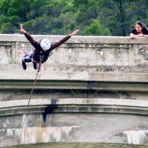 Alguien disfruta de "Puenting en Huesca: Vive la Caída Libre en Murillo de Gállego" desde un puente, mientras otra persona observa asombrada desde un costado. - ForTwo - Planes para parejas