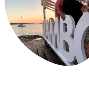 Un catamarán navega suavemente frente a la costa al atardecer con un cartel de MAMBO, que muestra la "Puesta de Sol en Catamarán en Ibiza: La Mejor Experiencia Todo Incluido". - ForTwo - Planes para parejas