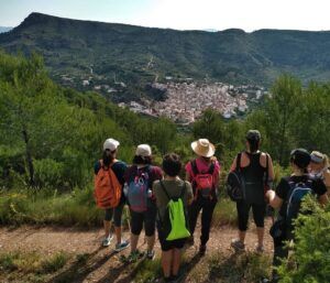 Los excursionistas con mochilas admiran un pueblo pintoresco y las exuberantes colinas verdes que aparecen en "Rutas de senderismo en Valencia: Naturaleza a tu ritmo". - ForTwo - Planes para parejas