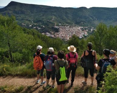 Los excursionistas con mochilas admiran un pueblo pintoresco y las exuberantes colinas verdes que aparecen en "Rutas de senderismo en Valencia: Naturaleza a tu ritmo". - ForTwo - Planes para parejas