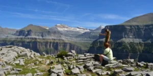 Dos personas admiran un vasto paisaje montañoso sobre un terreno rocoso bajo un cielo azul claro, perfecto para practicar senderismo con las "Rutas en 4x4 y Senderismo por el Parque Nacional de Ordesa. - ForTwo - Planes para parejas