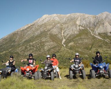 Los pilotos de cuatriciclos encarnan el espíritu aventurero de Rutas en Quad por la Alta Montaña de Huesca, posando ante una majestuosa Alta Montaña bajo un cielo azul despejado. - ForTwo - Planes para parejas