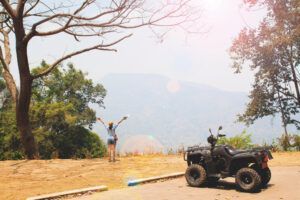 Cerca de un quad, una persona con los brazos en alto disfruta del impresionante paisaje de árboles y montañas de la Alta Montaña de Huesca durante una experiencia de Rutas en Quad por la Alta Montaña de Huesca. - ForTwo - Planes para parejas