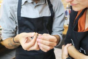 Dos personas con delantales inspeccionan atentamente un pequeño objeto, uno de ellos sosteniéndolo con cuidado. Se trata de un par del "Taller de Alianzas para Parejas en Barcelona: Crea tus Anillos a Mano", exquisitos y elaborados artesanalmente con precisión en Barcelona. - ForTwo - Planes para parejas
