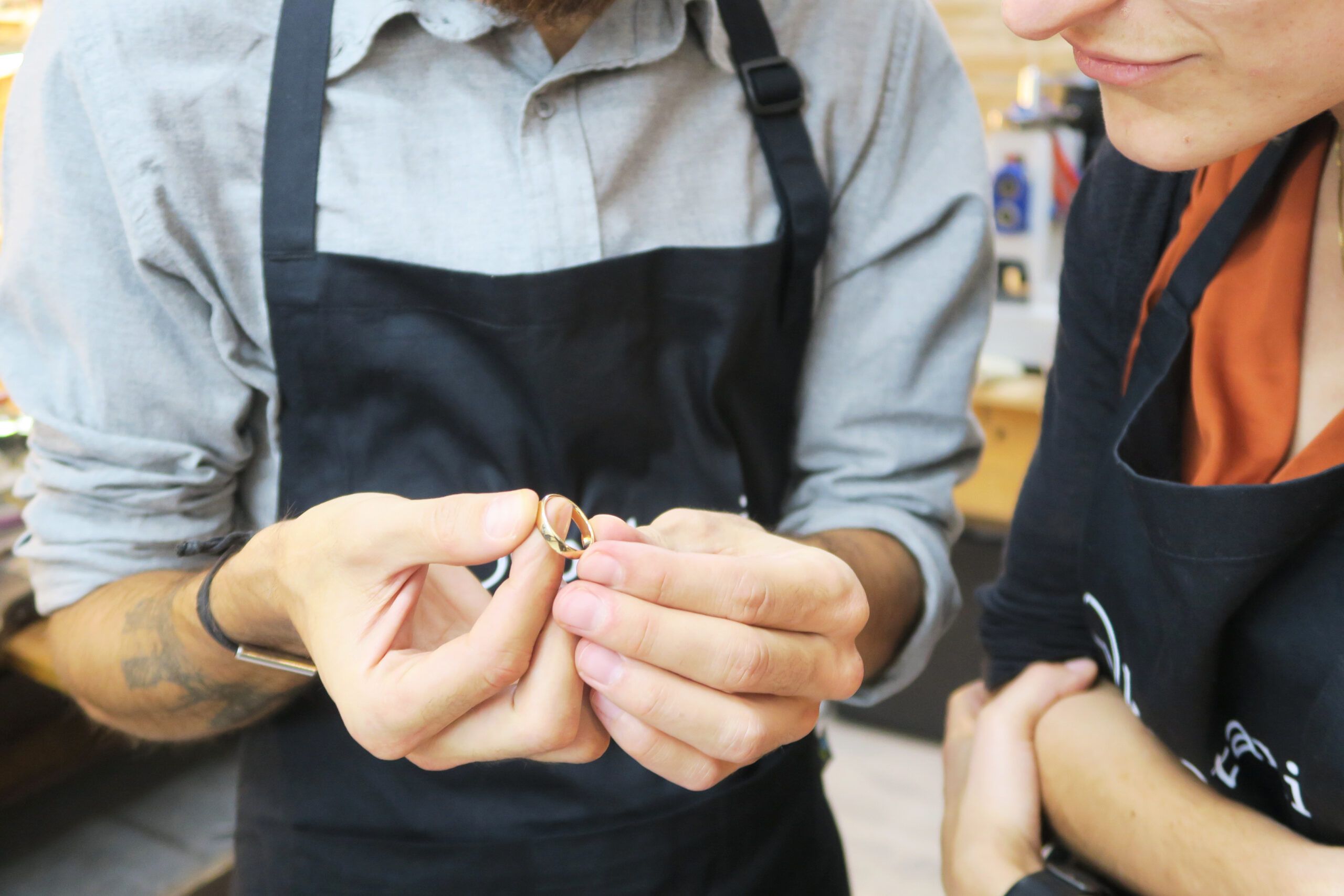 Dos personas con delantales inspeccionan atentamente un pequeño objeto, uno de ellos sosteniéndolo con cuidado. Se trata de un par del "Taller de Alianzas para Parejas en Barcelona: Crea tus Anillos a Mano", exquisitos y elaborados artesanalmente con precisión en Barcelona. - ForTwo - Planes para parejas