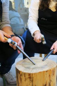 En un taller de Barcelona, dos personas colaboran en el "Taller de Alianzas para Parejas en Barcelona: Crea tus Anillos a Mano", utilizando hábilmente martillos sobre varillas de metal sobre un bloque de madera. - ForTwo - Planes para parejas