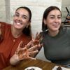 Dos mujeres sonríen con las manos cubiertas de chocolate en una mesa, disfrutando del "Taller de Cata y Elaboración de Chocolates Gourmet en Barcelona", deleitándose con deliciosos chocolates. - ForTwo - Planes para parejas