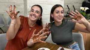 Dos mujeres sonríen con las manos cubiertas de chocolate en una mesa, disfrutando del "Taller de Cata y Elaboración de Chocolates Gourmet en Barcelona", deleitándose con deliciosos chocolates. - ForTwo - Planes para parejas