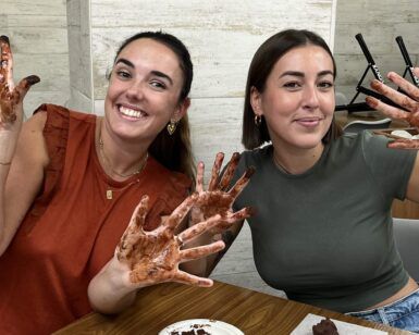 Dos mujeres sonríen con las manos cubiertas de chocolate en una mesa, disfrutando del "Taller de Cata y Elaboración de Chocolates Gourmet en Barcelona", deleitándose con deliciosos chocolates. - ForTwo - Planes para parejas
