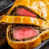 El Taller de Cocina para Sorprender a Tus Invitados ofrece una experiencia elegante y gourmet con filetes Wellington en rodajas sobre una tabla de madera, mostrando carne a medio cocinar envuelta en masa con una sabrosa capa de champiñones. - ForTwo - Planes para parejas
