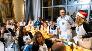 Un grupo animado con delantales, uno de ellos con un gorro de Papá Noel, se reúne alrededor de una mesa en el Taller de Cocina para Sorprender a Tus Invitados, sonriendo y charlando mientras se preparan para impresionar a sus invitados navideños. - ForTwo - Planes para parejas
