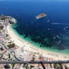 El "Tour en Helicóptero por Mallorca" ofrece una impresionante vista aérea de playas de arena, aguas turquesas con barcos que se mecen suavemente y una pequeña isla distante: una vista verdaderamente inolvidable. - ForTwo - Planes para parejas