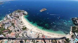 El "Tour en Helicóptero por Mallorca" ofrece una impresionante vista aérea de playas de arena, aguas turquesas con barcos que se mecen suavemente y una pequeña isla distante: una vista verdaderamente inolvidable. - ForTwo - Planes para parejas