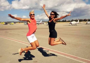 Dos mujeres saltan alegremente sobre un aeródromo con aviones y un cielo nublado soñando con su próxima emoción: un "Tour en Helicóptero por Mallorca" sobre las impresionantes vistas. - ForTwo - Planes para parejas