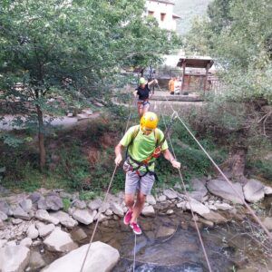 Con casco y arnés, una persona cruza un puente de cuerda sobre un arroyo, experimentando la emoción de la "Vía Ferrata en Huesca para Principiantes: Una Aventura Vertical entre Puentes y Escaleras" en los frondosos bosques de Huesca. - ForTwo - Planes para parejas