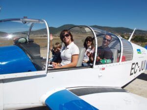 Una familia de cuatro personas disfruta de su vuelo en Avioneta sobre el Parque de Ordesa, sonriendo y saludando dentro de una pequeña avioneta en un día soleado con las pintorescas colinas del parque de fondo. - ForTwo - Planes para parejas